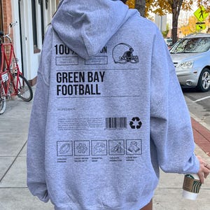 May include: A grey hoodie with the words "GREEN BAY FOOTBALL" and a football helmet graphic. The back of the hoodie features a barcode, recycling symbol, and icons representing football themes. The hoodie is made of a soft material.