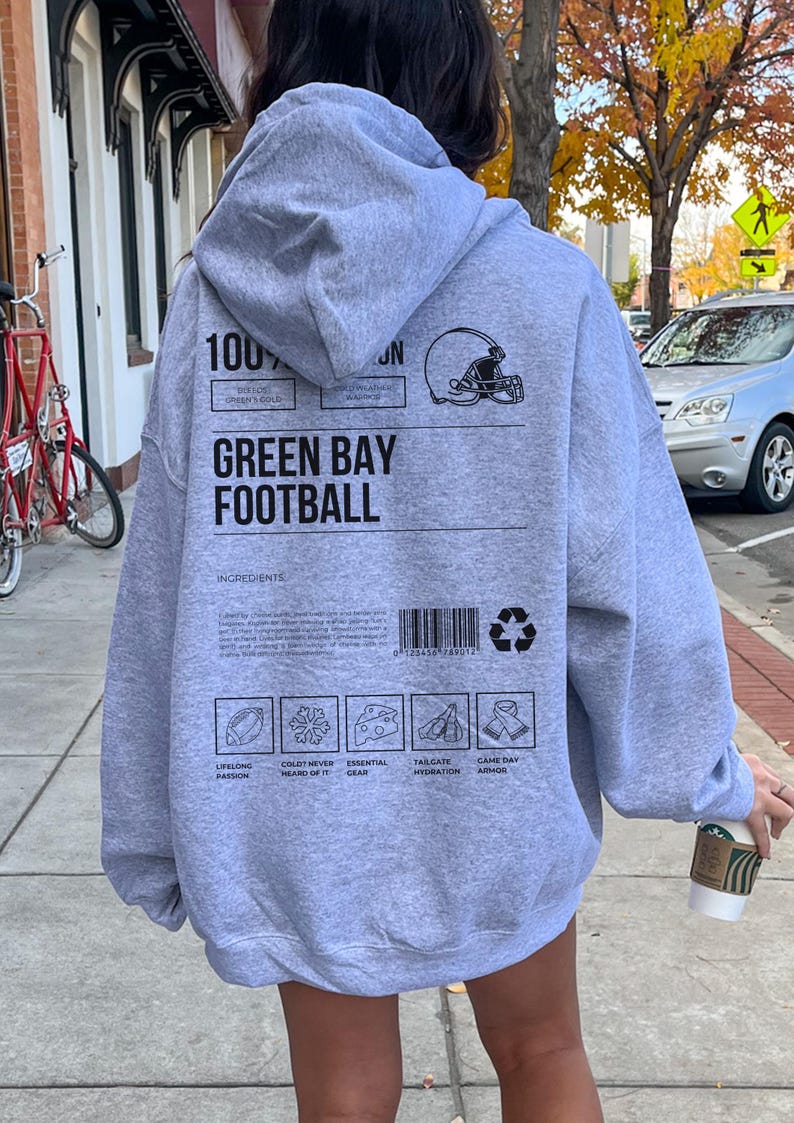 May include: A grey hoodie with the words "GREEN BAY FOOTBALL" and a football helmet graphic. The back of the hoodie features a barcode, recycling symbol, and icons representing football themes. The hoodie is made of a soft material.