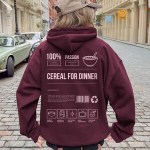 Może przedstawiać: Bordowa bluza z kapturem z napisem "CEREAL FOR DINNER" i elementami graficznymi. Tył bluzy z kapturem zawiera słowa "100% PASSION" i "INGREDIENTS". Osoba ma na sobie beżową czapkę z daszkiem.