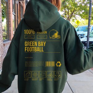May include: Dark green hoodie with gold text and graphics. The back of the hoodie features the words "GREEN BAY FOOTBALL" and "100% PASSION". Additional text includes "BLEEDS GREEN & COLD" and "COLD WEATHER WARRIOR".
