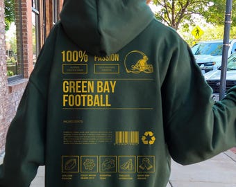 May include: Dark green hoodie with gold text and graphics. The back of the hoodie features the words "GREEN BAY FOOTBALL" and "100% PASSION". Additional text includes "BLEEDS GREEN & COLD" and "COLD WEATHER WARRIOR".