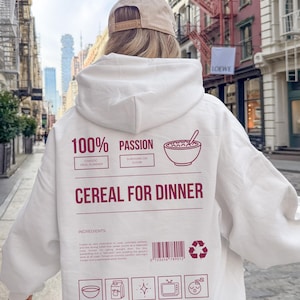 Może przedstawiać: Biała bluza z kapturem z bordowym napisem "100% Passion" i "Cereal for Dinner". Projekt zawiera grafikę miski z płatkami śniadaniowymi, listę składnik&oacute;w i ikony. Noszona jest beżowa czapka.