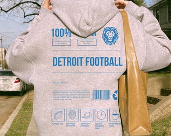 Peut inclure: Un sweat à capuche gris clair avec le texte "DETROIT FOOTBALL" en bleu. Le dos du sweat à capuche présente un logo de lion bleu et diverses icônes. Le sweat à capuche a une capuche à cordon et des manches longues. Un sac fourre-tout marron est porté sur l'épaule.