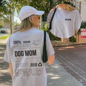 May include: Gray t-shirt with the text "DOG MOM" on the front and a graphic on the back that says "100% Passion Dog Mom" with a list of ingredients: "A person who treats their dog better than themselves.  You can find them walking their dog, playing fetch, and caring for their dog's needs.  They handle vet visits and other stressful situations with grace and patience."