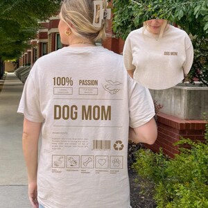 May include: A beige t-shirt with the text "DOG MOM" printed on the front and a graphic on the back that says "100% Passion" with a list of ingredients for being a dog mom, including "walk enthusiast", "treat dispenser", "unconditional love", and "pawsitive parent".