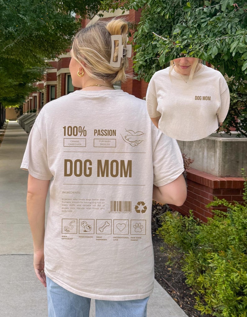 May include: A beige t-shirt with the text "DOG MOM" printed on the front and a graphic on the back that says "100% Passion" with a list of ingredients for being a dog mom, including "walk enthusiast", "treat dispenser", "unconditional love", and "pawsitive parent".