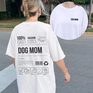 May include: White t-shirt with a graphic print on the back that says "100% Passion" and "Dog Mom". The graphic includes a dog paw print and a heart. The front of the shirt has the text "Dog Mom" printed on it.