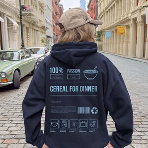 Może przedstawiać: Granatowa bluza z kapturem z napisem "100% Passion Cereal for Dinner". Tył bluzy zdobi grafika miski z płatkami i ikony składnik&oacute;w. Osoba ma na sobie beżową czapkę z daszkiem.