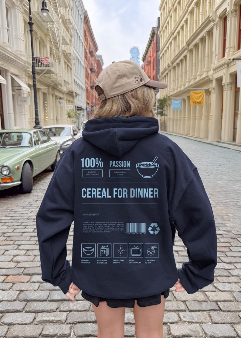 Może przedstawiać: Granatowa bluza z kapturem z napisem "100% Passion Cereal for Dinner". Tył bluzy zdobi grafika miski z płatkami i ikony składnik&oacute;w. Osoba ma na sobie beżową czapkę z daszkiem.