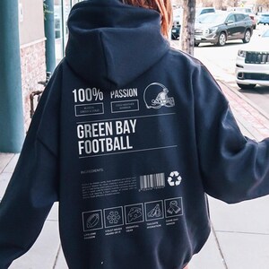May include: Dark navy blue hoodie with white text and graphics. The back of the hoodie reads "100% PASSION" and "GREEN BAY FOOTBALL". Includes a football helmet graphic and ingredient icons. A barcode and recycling symbol are also present.