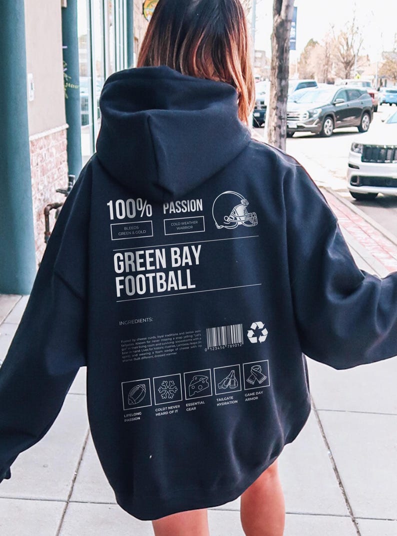 May include: Dark navy blue hoodie with white text and graphics. The back of the hoodie reads "100% PASSION" and "GREEN BAY FOOTBALL". Includes a football helmet graphic and ingredient icons. A barcode and recycling symbol are also present.