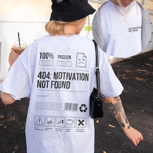 Könnte beinhalten: Hellgraues T-Shirt mit dem schwarzen Text "404: MOTIVATION NOT FOUND". Das Shirt zeigt auch verschiedene Symbole und die Worte "100% PASSION". Ein schwarzer Fischerhut und eine schwarze Handtasche sind ebenfalls zu sehen.