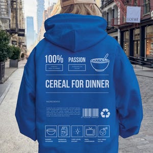 Może przedstawiać: Niebieska bluza z kapturem z białym napisem "100% PASSION" i "CEREAL FOR DINNER". Z tyłu znajduje się grafika miski z płatkami śniadaniowymi i inne ikony. Bluza jest noszona przez osobę w środowisku miejskim.