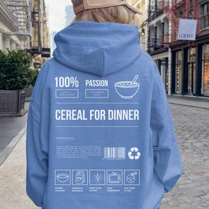 Może przedstawiać: Jasnoniebieska bluza z kapturem z białym napisem "CEREAL FOR DINNER". Tył bluzy z kapturem zawiera r&oacute;wnież słowa "100% PASSION" i r&oacute;żne ikony. Osoba ma na sobie beżową czapkę z daszkiem.
