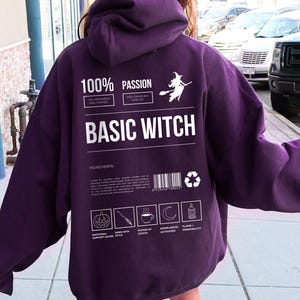 Könnte beinhalten: Ein dunkelvioletter Kapuzenpullover mit dem weißen Text "BASIC WITCH". Auf der Rückseite des Kapuzenpullovers befindet sich eine Hexe auf einem Besen und andere Grafiken. Der Kapuzenpullover hat eine Kapuze und lange Ärmel.