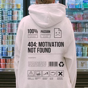 Op de afbeelding: Een lichtgrijze hoodie met de tekst "100% Passion" en "404: Motivation Not Found" op de rug. Het ontwerp bevat iconen en tekst over werk en motivatie. De hoodie heeft een capuchon en een buidelzak.