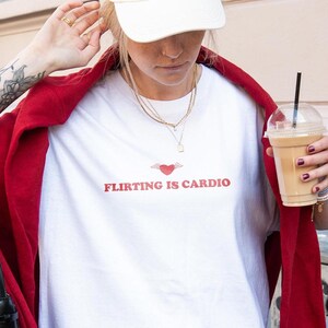 Op de afbeelding: Wit T-shirt met de rode tekst "Flirting is Cardio", met een hart met vleugels. De persoon draagt een witte pet, gouden kettingen en een rode jas. Ze houdt een koffiedrank vast.