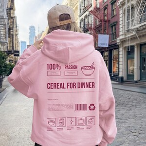 Może przedstawiać: Jasnor&oacute;żowa bluza z kapturem z napisem "100% Passion Cereal for Dinner" z tyłu. Bluza ma kaptur i długie rękawy. Tło przedstawia ulicę miasta z budynkami.