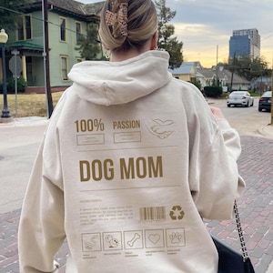 May include: Beige hoodie with a brown graphic print that says "100% Passion Canine Caregiver Fur Baby Spoiler Dog Mom". The graphic also includes a heart, paw print, bone, and a ball. The graphic is labeled "Ingredients" and includes a list of items: "Walk Enthusiast, Fetch Fanatic, Treat Dispenser, Unconditional Love, Paw-some Parent."