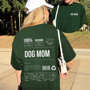 May include: Green t-shirt with white text that reads "100% Passion Canine Caregiver Fur Baby Spoiler Dog Mom". The shirt also has a graphic of a hand holding a paw. The text "Ingredients" is followed by a description of a dog mom. The shirt also has a barcode and recycling symbol. Below the description are five icons: Walk Enthusiast, Fetch Fanatic, Treat Dispenser, Unconditional Love, Paw-some Parent.