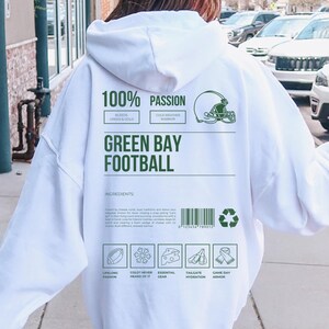 May include: White hoodie with green text reading "100% PASSION" and "GREEN BAY FOOTBALL". The back of the hoodie features a football helmet graphic and ingredient icons. A casual, comfortable garment.
