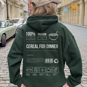 Może przedstawiać: Ciemnozielona bluza z kapturem z napisem "CEREAL FOR DINNER" oraz innymi tekstami i grafikami. Bluza z kapturem ma kaptur i długie rękawy. Tekst zawiera "100% PASSION" i "INGREDIENTS".