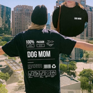 May include: Black t-shirt with a white graphic design that says "100% Passion" and "Dog Mom". The graphic design is a label with a barcode and recycling symbol. The label also includes icons for "Wolf Enthusiast", "Fetch Fanatic", "Treat Obsessed", "Unconditional Love", and "Ever-Loving Parent".