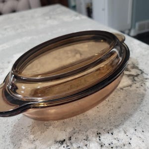 May include: An oval-shaped, amber-colored glass casserole dish with a matching lid. The dish has a rounded base and a slightly flared rim. The lid fits snugly on top, and the dish has two small handles on either side.