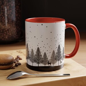 May include: A white ceramic mug with a red rim and handle. The mug features a black and white design of a snowy forest with trees and deer. The mug is sitting on a wooden surface.
