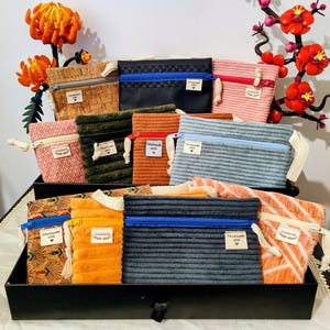 Può includere: Una collezione di pochette con cerniera fatte a mano in vari colori e texture, tra cui velluto a coste, sughero e tessuti fantasia. Ogni pochette ha una piccola etichetta con la scritta "handmade with love". Le pochette sono esposte in un vassoio nero, con fiori decorativi sullo sfondo.