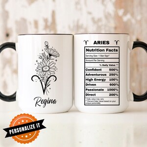 May include: White ceramic mug with a black handle and rim. One side features a floral design with the name "Regina" below. The other side displays "Aries Nutrition Facts" with personality traits and percentages. A "Personalize It" sticker is at the bottom.