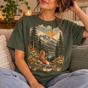 Peut inclure: Un t-shirt vert olive foncé avec une impression graphique représentant un renard assis sur un rocher, surplombant un lac, des pins et des montagnes enneigées sous un soleil jaune, le tout encadré par un motif texturé de planches de bois.