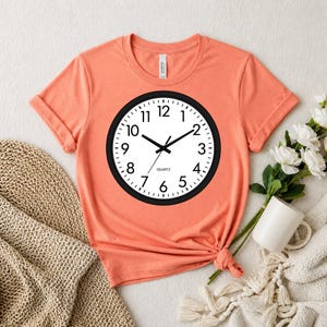 May include: A coral-colored short-sleeve t-shirt featuring a large graphic of a round wall clock with a black frame, a white face, black numerals, black hands, and the word QUARTZ printed in black text near the center of the clock face.
