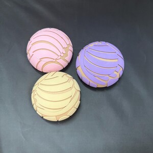 May include: Three colourful, round, decorative items with a unique design. The items are pink, purple, and yellow with a pattern of lines and curves.