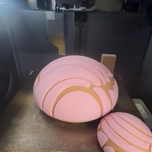May include: A 3D printer is shown with two pink concha bread shapes in the process of being printed. The conchas have a light brown, textured design on top. The printer is gray and white.