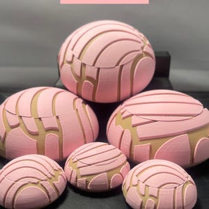 May include: A collection of six 3D printed concha bread rolls in various sizes. The rolls are pink and brown with a detailed, textured surface. The rolls are arranged in a pyramid shape on a black surface.