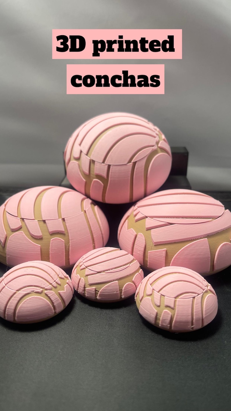 May include: A collection of six 3D printed concha bread rolls in various sizes. The rolls are pink and brown with a detailed, textured surface. The rolls are arranged in a pyramid shape on a black surface.