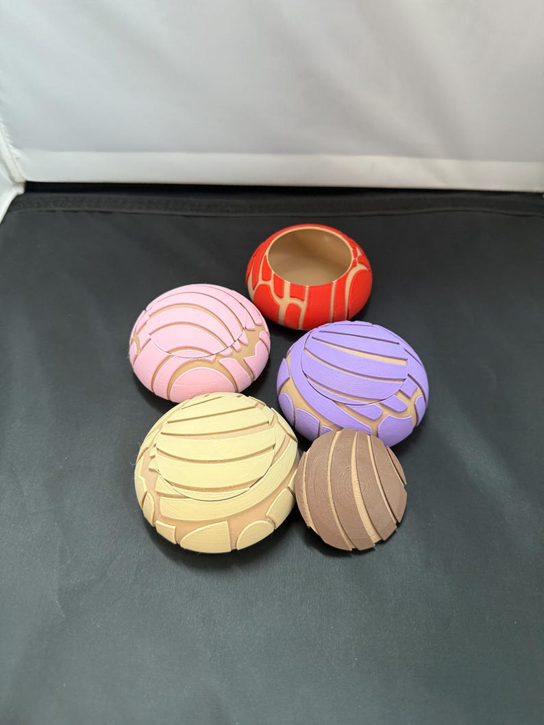 May include: Five colourful ceramic bowls shaped like Mexican sweet bread, known as conchas. The bowls are in various colours, including pink, purple, brown, beige, and red.