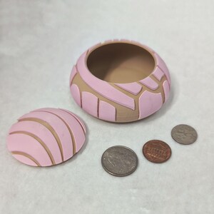 May include: A pink and brown ceramic container with a lid. The container is shaped like a traditional Mexican pastry called a concha. The lid is separate and has a similar design to the container.