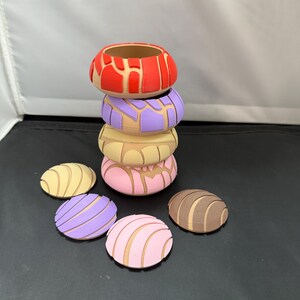 May include: A stack of six colourful, round, decorative ceramic concha shells. The shells are in various colours, including red, purple, pink, brown, and yellow. The shells are stacked on top of each other, with the largest shell at the bottom and the smallest shell at the top.