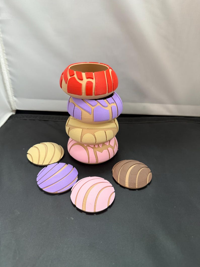 May include: A stack of six colourful, round, decorative ceramic concha shells. The shells are in various colours, including red, purple, pink, brown, and yellow. The shells are stacked on top of each other, with the largest shell at the bottom and the smallest shell at the top.