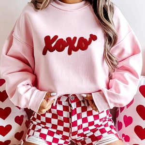 May include: A pink sweatshirt with the word "XOXO" embroidered in red on the front. The sweatshirt is worn over a red and white checkered skirt.