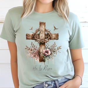 May include: A light green t-shirt featuring a watercolor design of a wooden cross with a crown of thorns, flowers, and two birds. The text "He Is Risen" is printed below the cross.