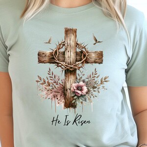 May include: A light blue t-shirt featuring a wooden cross with a crown of thorns, floral accents, and two flying birds. The text "He Is Risen" is printed below the cross. The design is centered on the shirt.