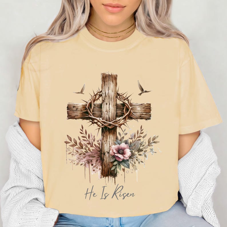 May include: A light yellow t-shirt featuring a watercolor design of a wooden cross with a crown of thorns, flowers, and two birds. The text "He Is Risen" is printed below the cross. The shirt has short sleeves.