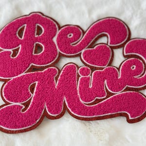 Puede incluir: Parche de fieltro rosa con las palabras "Be Mine" en un estilo de fuente retro. Las letras tienen un contorno blanco y un corazón está incorporado en la "M".
