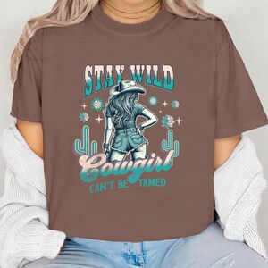 May include: Brown t-shirt featuring a graphic of a woman wearing a cowboy hat and jeans. The text "Stay Wild Cowgirl Can't Be Tamed" is printed on the shirt.