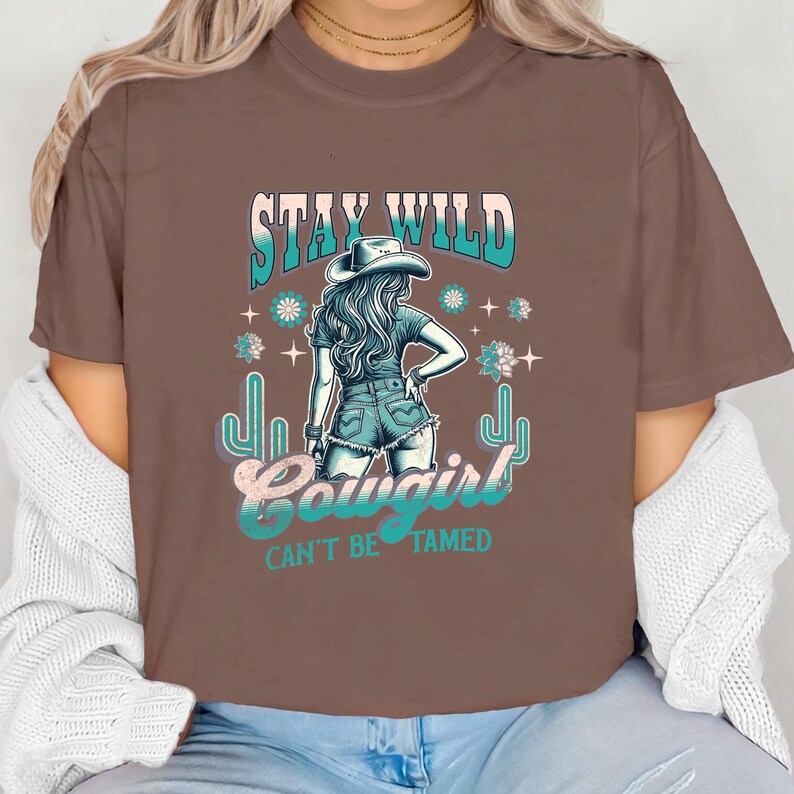 May include: Brown t-shirt featuring a graphic of a woman wearing a cowboy hat and jeans. The text "Stay Wild Cowgirl Can't Be Tamed" is printed on the shirt.