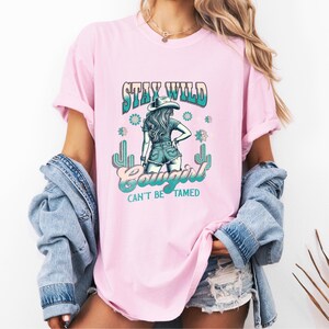 May include: Pink t-shirt featuring a graphic of a cowgirl wearing a hat and jeans. The text "Stay Wild Cowgirl Can't Be Tamed" is printed on the shirt.