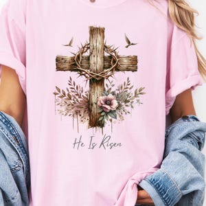 May include: Pink t-shirt featuring a watercolor design of a wooden cross with a crown of thorns, floral accents, and two birds. The text "He Is Risen" is printed below the cross.